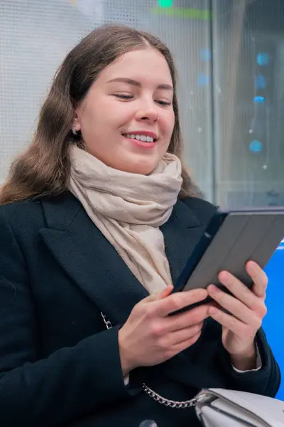 Modern bir metro treninde oturan siyah paltolu ve bej eşarplı odaklanmış genç bir kadın bir e-okuyucuda okuyor. Her gün işe gidip gelirken sakin bir anın tadını çıkarıyor.. 