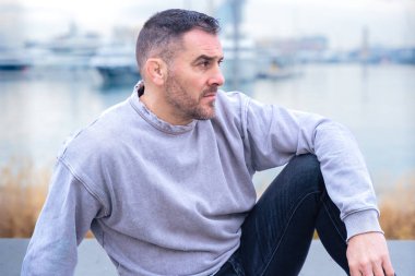 Gri bir kazak giyen rahat bir adam marinaya yakın bir yerde oturuyor ve kameraya güvenle bakıyor. Sakin ortam ve hafif ışıklandırma onun düşünceli yüz ifadesini güçlendiriyor.. 