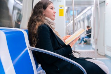 Siyah paltolu ve bej eşarplı genç bir kadın modern bir metro treninde oturmuş, odaklanmış bir kitap okuyor. Şehirdeki seyahati sırasında sakin bir anın tadını çıkarıyor.. 