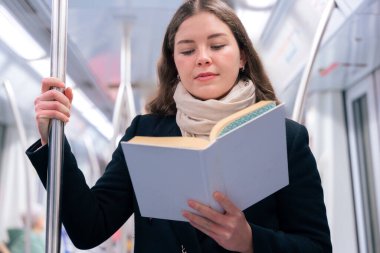 Modern bir metro treninde duran siyah paltolu ve bej eşarplı odaklanmış genç bir kadın metal bir direğe tutunarak kitap okuyor. Seyahat sırasında sakin bir anın tadını çıkarıyor..