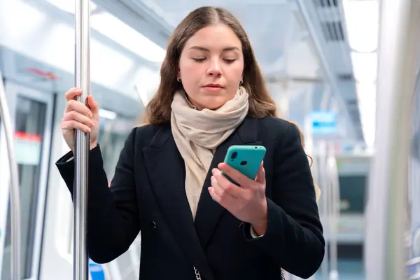 Siyah paltolu ve bej eşarplı genç bir kadın modern bir metro treninin içinde elinde akıllı bir telefonla metal bir direği kavrıyor. İşe gidip gelirken odaklanmış görünüyor. Elinde şık beyaz bir çanta var.. 