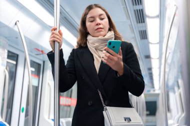 Siyah paltolu ve bej eşarplı genç bir kadın modern bir metro treninin içinde elinde akıllı bir telefonla metal bir direği kavrıyor. İşe gidip gelirken odaklanmış görünüyor. Elinde şık beyaz bir çanta var.. 