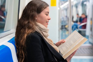 Siyah paltolu ve bej eşarplı genç bir kadın modern bir metro treninde oturmuş, odaklanmış bir kitap okuyor. Şehirdeki seyahati sırasında sakin bir anın tadını çıkarıyor.. 