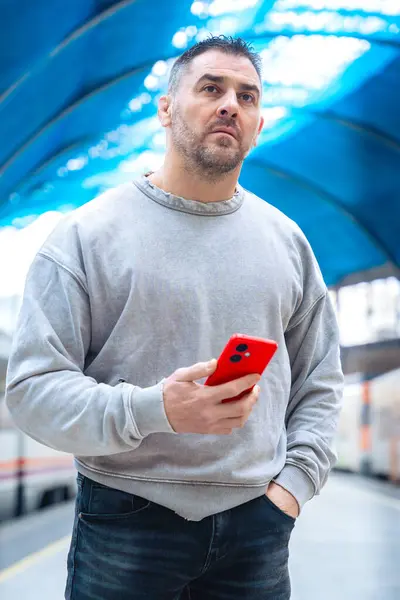  Tren istasyonuna bakarken elinde kırmızı bir akıllı telefon tutan gri kazak giyen ciddi bir adam. İfadesi beklenti, seyahat ve modern bağlılık taşıyor..
