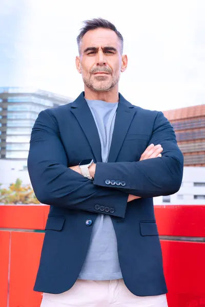 Şık takım elbiseli orta yaşlı bir işadamı kollarını kavuşturmuş, güven ve liderlik yayıyor. Arka plandaki modern şehir manzarası profesyonel ve otoriter görünümü geliştirir.. 