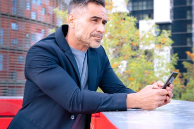 Orta yaşlı bir işadamı şık bir takım elbiseyle korkuluklara dayanıyor, akıllı telefonunu odaklanmış bir ifadeyle tutuyor. Modern şehir arkaplanı profesyonel ve düşünceli bir ruh halini yansıtır.. 