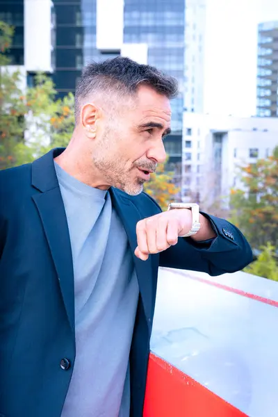 Takım elbiseli orta yaşlı bir işadamı akıllı bakışlarını odaklanmış bir ifadeyle kontrol ediyor. Modern şehir arkaplanı profesyonel ve zamana duyarlı bir yaşam tarzını vurgular.