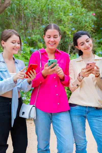 Dışarıda duran üç genç kadın akıllı telefonlarına bakıp gülümsüyor. Konuşmaktan ya da içeriği paylaşmaktan zevk alıyor gibi görünüyorlar. Günlük giysiler ve yeşil arka plan.