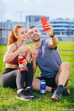 Gülümseyen genç adam ve kadın çimenlerde oturup egzersiz yaptıktan sonra selfie çekiyorlar. Spor kıyafetleri giyiyorlar ve ellerinde su şişeleri var. Şehir parkında rahat bir anın tadını çıkarıyorlar..