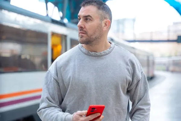  Tren istasyonuna bakarken elinde kırmızı bir akıllı telefon tutan gri kazak giyen ciddi bir adam. İfadesi beklenti, seyahat ve modern bağlılık taşıyor..