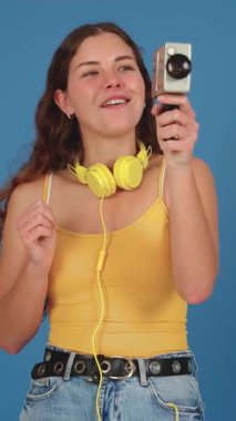  Sarı kulaklıklı ve sarı bluzlu gülümseyen genç bir kadın fotoğraf makinesiyle fotoğraf çekiyor. Heyecanlı ve neşeli görünüyor, saf mavi bir arka planda duruyor, mutluluğu ifade ediyor..