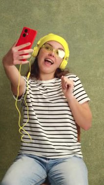 Kulaklıklı gülümseyen genç bir kadın evde otururken kırmızı bir akıllı telefonla selfie çekiyor. Çizgili bir tişört giyiyor ve şakacı poz veriyor. Yeşil bitkiler ve arka planda desenli bir duvar..