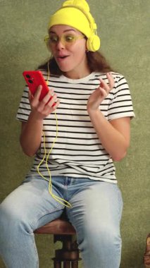 Sarı bir bere, kulaklık ve çizgili gömlek takan neşeli genç bir kadın kırmızı akıllı telefonunu tutarken heyecanla kutluyor. Bitkilerin yeşil arka planı yeni bir atmosfer ekler.. 