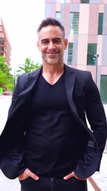  Siyahlar giymiş şık bir adam, şehir ortamında dikilirken kendine güvenerek gülümsüyor. Arka plandaki modern mimari profesyonel ve karizmatik varlığa katkı sağlar. 