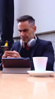 Kafede akıllı telefonunu kullanırken kameraya gülümseyen kulaklıklı olgun bir adam. Masanın üzerinde bir kahve fincanı ve tablet, modern ve profesyonel bir ortam yaratıyor.. 