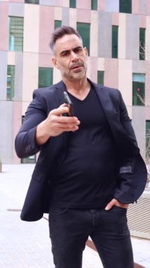 Siyah kıyafetli şık bir adam akıllı telefonu tutarken başını sallıyor. Arka planda modern bir binayla kentsel bir ortamda durmak, anlaşmazlığı ifade etmek veya reddetmek. 