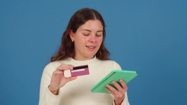 Elinde kredi kartı ve tablet tutan genç bir kadın, düşünceli görünüyor. Beyaz bir kazak giyiyor ve mavi bir arka plana karşı duruyor. Çevrimiçi alışveriş, ödeme veya mali kararlar kavramı.