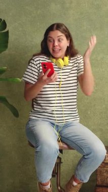 Sarı kulaklıklı ve kırmızı akıllı telefonlu genç bir kadın evde otururken neşesini gösteriyor. Çizgili bir tişört giyiyor ve ışıl ışıl gülümsüyor. Yeşil bitkiler ve arka planda desenli bir duvar.. 