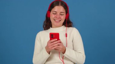 Kırmızı kulaklık takan, elinde akıllı bir telefonla dans eden ve en sevdiği müziğin keyfini çıkaran neşeli genç bir kadın. Mavi arka planda neşe ve enerjiyi ifade etmek. 