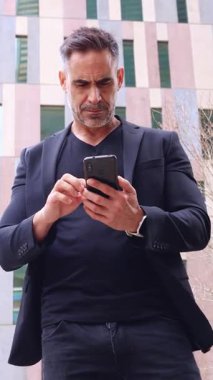 Siyah kıyafetli mutlu adam akıllı telefonuna bakarken başarıyı kutluyor. Neşeli ifadesi ve sıkılmış yumruğu şehir ortamında heyecan ve başarı taşıyor..