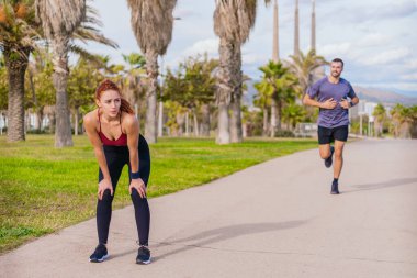 Bir adam palmiye ağaçlarıyla parkta koşmaya devam ederken genç bir kadın koşudan sonra soluklanıyor. Her ikisi de spor giyer ve kıyıya yakın güneşli bir günde dışarıda egzersiz yaparlar.. 