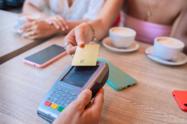  Bir kadının kredi kartıyla bir kafede temassız ödeme yaptığı yakın plan fotoğraf. Kahve fincanları, akıllı telefonlar ve arka planda modern kolaylığı sembolize eden rahat bir atmosfer..