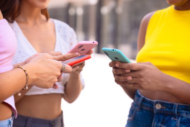 Üç neşeli genç kadın kırmızı bir akıllı telefonun etrafında toplanıyor, heyecanla tepki veriyorlar. Gelişigüzel giyiniyorlar, modern bir şehir ortamında dışarıda duruyorlar, birlikte eğlenceli bir anın tadını çıkarıyorlar..