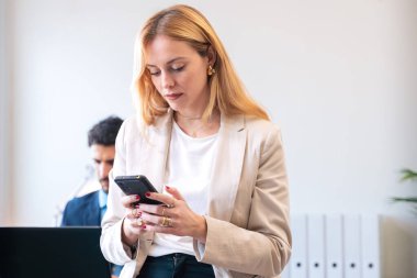  Takım elbiseli profesyonel bir kadın parlak bir ofiste akıllı telefonuna konsantre oluyor. Arka planda çalışan bir bayan meslektaşım, ortak bir çalışma ortamı yaratıyor..