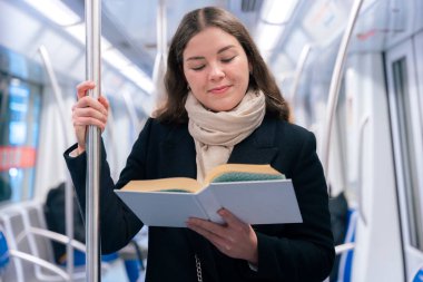Modern bir metro treninde duran siyah paltolu ve bej eşarplı odaklanmış genç bir kadın metal bir direğe tutunarak kitap okuyor. Seyahat sırasında sakin bir anın tadını çıkarıyor..