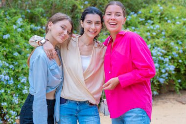 Üç genç kadın, yemyeşil bir parkta poz verirken gülümsüyor ve birbirlerine sarılıyorlar. Havalı günlük kıyafetler giyiyorlar, mutluluğu, arkadaşlığı ve eğlenceyi açık havada ifade ediyorlar.. 