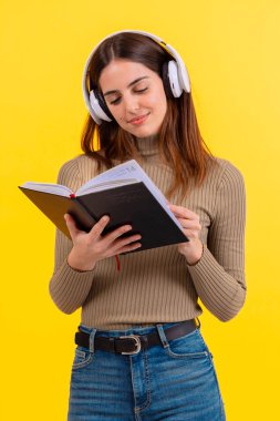 Beyaz kulaklık takan, elinde açık bir kitapla, parlak sarı bir arka plana karşı duran neşeli genç bir kadın. Öğrenme, işitme ve eğitim konuları için mükemmel bir resim. Dikey