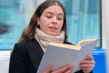Siyah paltolu ve bej eşarplı genç bir kadın modern bir metro treninde oturmuş, odaklanmış bir kitap okuyor. Şehirdeki seyahati sırasında sakin bir anın tadını çıkarıyor.. 