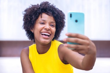  Sarı bluzlu, kıvırcık saçlı neşeli genç bir kadın açık havada selfie çekiyor, gülümsüyor ve barış işareti yapıyor. Canlı sahne mutluluk, bireysellik ve modern yaşam tarzını gösteriyor..