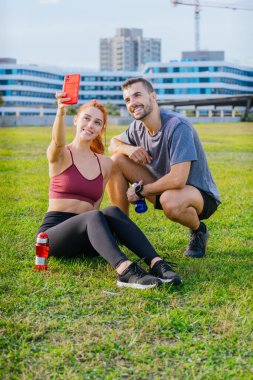 Gülümseyen genç adam ve kadın çimenlerde oturup egzersiz yaptıktan sonra selfie çekiyorlar. Spor kıyafetleri giyiyorlar ve ellerinde su şişeleri var. Şehir parkında rahat bir anın tadını çıkarıyorlar..