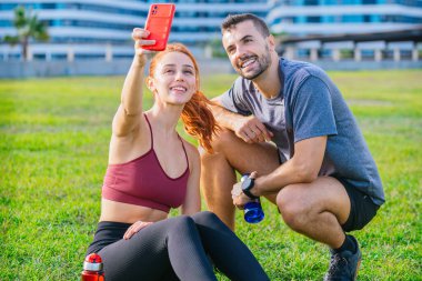 Gülümseyen genç adam ve kadın çimenlerde oturup egzersiz yaptıktan sonra selfie çekiyorlar. Spor kıyafetleri giyiyorlar ve ellerinde su şişeleri var. Şehir parkında rahat bir anın tadını çıkarıyorlar..