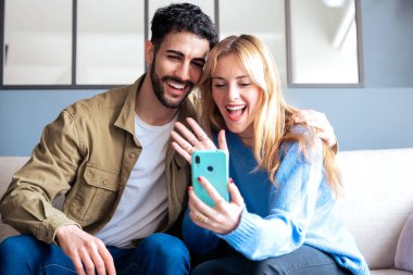 Neşeli bir çift rahat bir kanepede oturur ve görüntülü konuşma sırasında gülümser ve akıllı telefona el sallarlar. Onların mutlu ifadeleri ve rahatlamış ortam bağlantıları, teknolojileri ve ilişkileri.