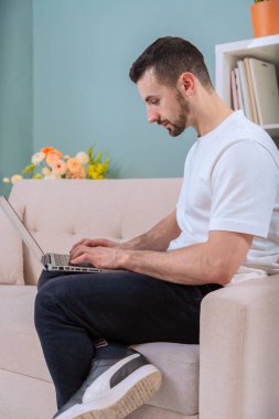  Bir adam rahat bir oturma odasında rahat bir kanepeye oturur ve dizüstü bilgisayarıyla çalışır. Odada bitkiler, bir ayna ve yumuşak ışıklandırma var. Üretkenlik için sıcak ve huzurlu bir ortam yaratıyorlar..