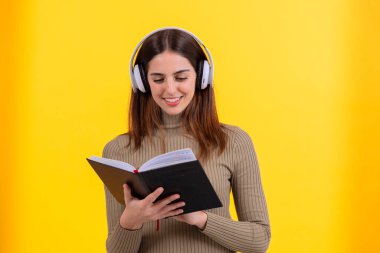 Beyaz kulaklık takan, elinde açık bir kitapla, parlak sarı bir arka plana karşı duran neşeli genç bir kadın. Öğrenme, sesli kitap ya da eğitim temaları için mükemmel bir resim.
