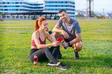 Yeşil bir parkta spor giyimli genç bir çift egzersizden sonra dinleniyor. Kadın akıllı telefonunu kullanarak çimlerin üzerinde otururken, adam elinde su şişesiyle çömeliyor, ikisi de gülümsüyor.. 