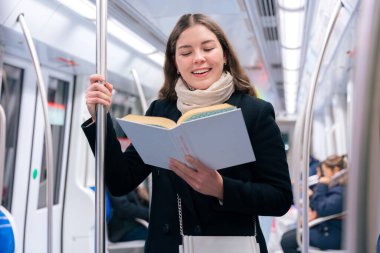 Modern bir metro treninde duran siyah paltolu ve bej eşarplı odaklanmış genç bir kadın metal bir direğe tutunarak kitap okuyor. Seyahat sırasında sakin bir anın tadını çıkarıyor..