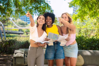 Üç neşeli genç kadın, elinde haritalar ve bir şeye işaret ederken bir seyahat rehberi tutarak dışarıda duruyorlar. Heyecanla gülümserler, güneşli bir şehir ortamında ağaçlar ve binalarla maceralarının tadını çıkarırlar.