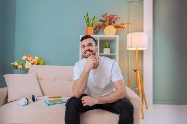 Bir adam derin düşüncelere dalmış, rahat bir oturma odasında oturuyor. Kanepede, çenesinde bir eliyle, bitkilerle ve hafif ışıklandırmayla rahatlamış, yansıması için huzurlu bir atmosfer yaratıyor..