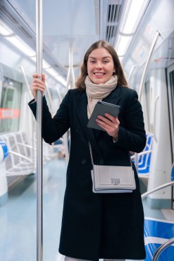 Modern bir metro treninde dururken siyah paltolu ve bej eşarplı şık bir kadın bir e-okuyucuda okuyor. Yol boyunca bir direğe tutunarak kameraya bakarak dengede duruyor.
