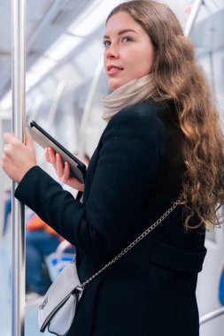 Modern bir metro treninde dururken siyah paltolu ve bej eşarplı şık bir kadın bir e-okuyucuda okuyor. Yolda giderken bir direğe tutunarak dengede duruyor.. 