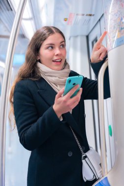  Siyah ceketli ve bej eşarplı odaklanmış genç bir kadın akıllı telefonunu tutarken metro haritasını kontrol ediyor. Modern bir metro treninde rotasını çiziyor..