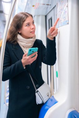 Siyah ceketli ve bej eşarplı odaklanmış genç bir kadın akıllı telefonunu tutarken metro haritasını kontrol ediyor. Modern bir metro treninde rotasını çiziyor..