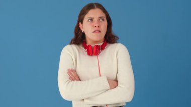 Boynuna kırmızı kulaklık takan genç bir kadın kafa karışıklığı ve hayal kırıklığını ifade ediyor. Şaşırmış görünürken tek eliyle hareket ediyor. Düz mavi arkaplan. 