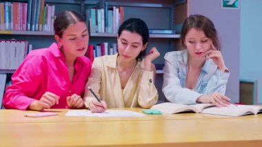 Kütüphanede bir masada oturan üç genç kadın önlerinde kitaplar, kağıtlar ve bir telefon olan görevler üzerinde birlikte çalışıyorlar..