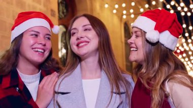 Üç mutlu arkadaş Noel 'i kutluyor, Noel Baba şapkaları takıyor ve festival ışıklarının keyfini çıkarıyorlar. Arka planda parlak tatil dekorasyonlarıyla birlikte dışarıda poz veriyorlar, neşe saçıyorlar..