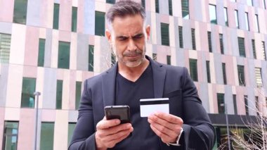 Elinde kredi kartı ve akıllı telefon olan siyah elbiseli bir adam online ödeme yapıyor. Arka planda modern binaların bulunduğu kentsel bir ortamda dijital işlemleri temsil ediyor.. 